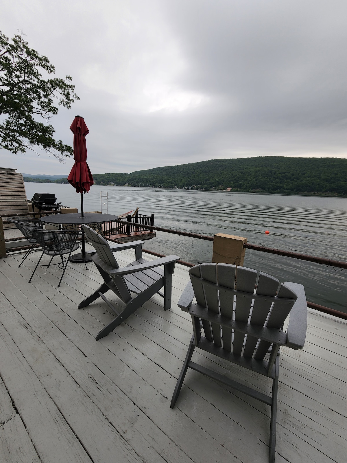 A spacious waterfront deck features several seating options including two adirondack chairs and a dining table with chairs. A large umbrella provides shade. The calm waters of the lake reflect the surrounding green hills under a cloudy sky.