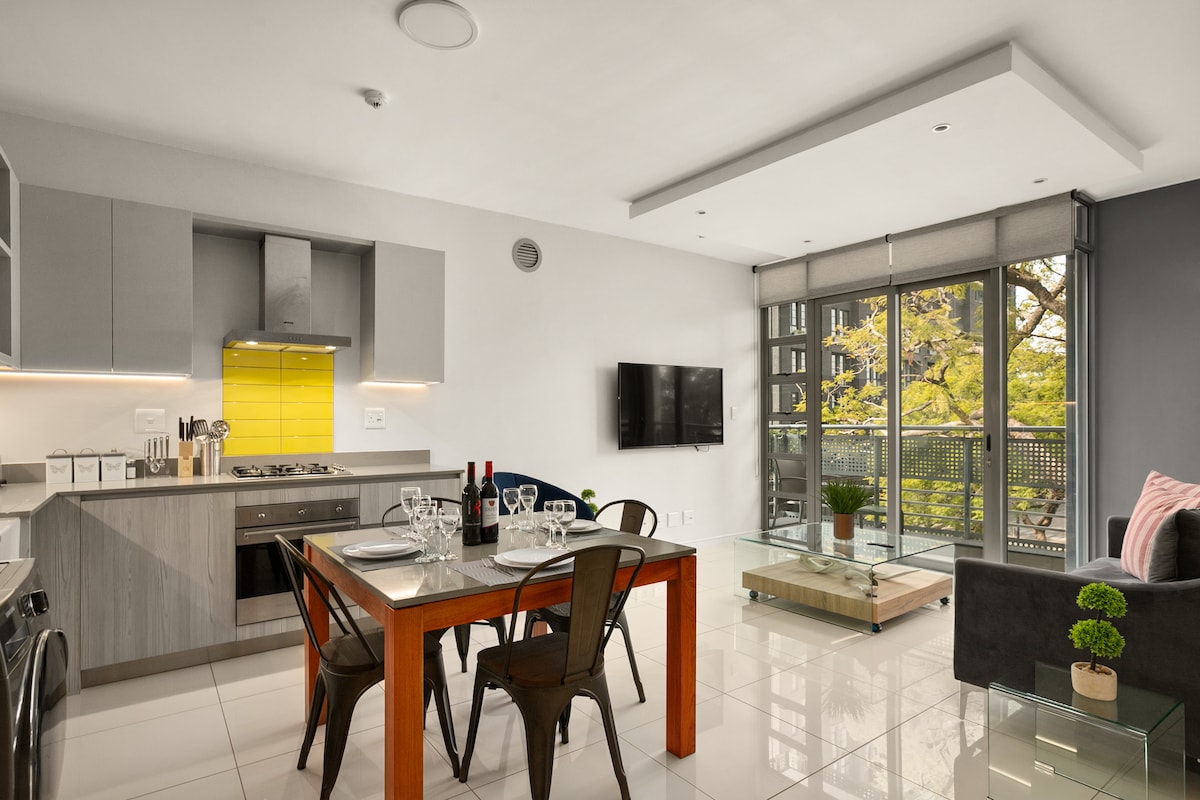 A modern kitchen and dining area are featured, showcasing a wooden dining table set for four. Clean lines and minimalist design characterize the space, with a sleek kitchen highlighted by gray cabinets and a bright yellow backsplash. Large windows offer views of greenery outside.