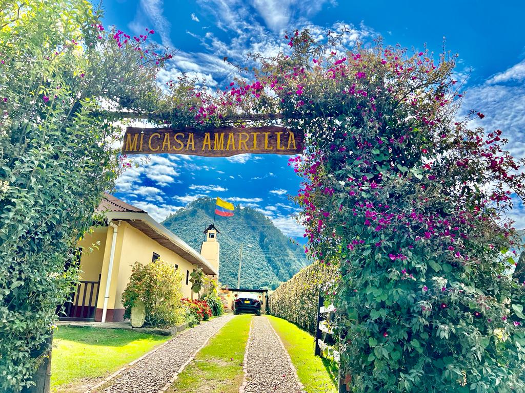 A welcoming entrance is framed by vibrant flowering plants and a sign reading 'Mi Casa Amarilla.' Lush greenery lines the pathway leading to the house, with a mountainous backdrop under a bright blue sky. The Colombian flag is visible in the distance.