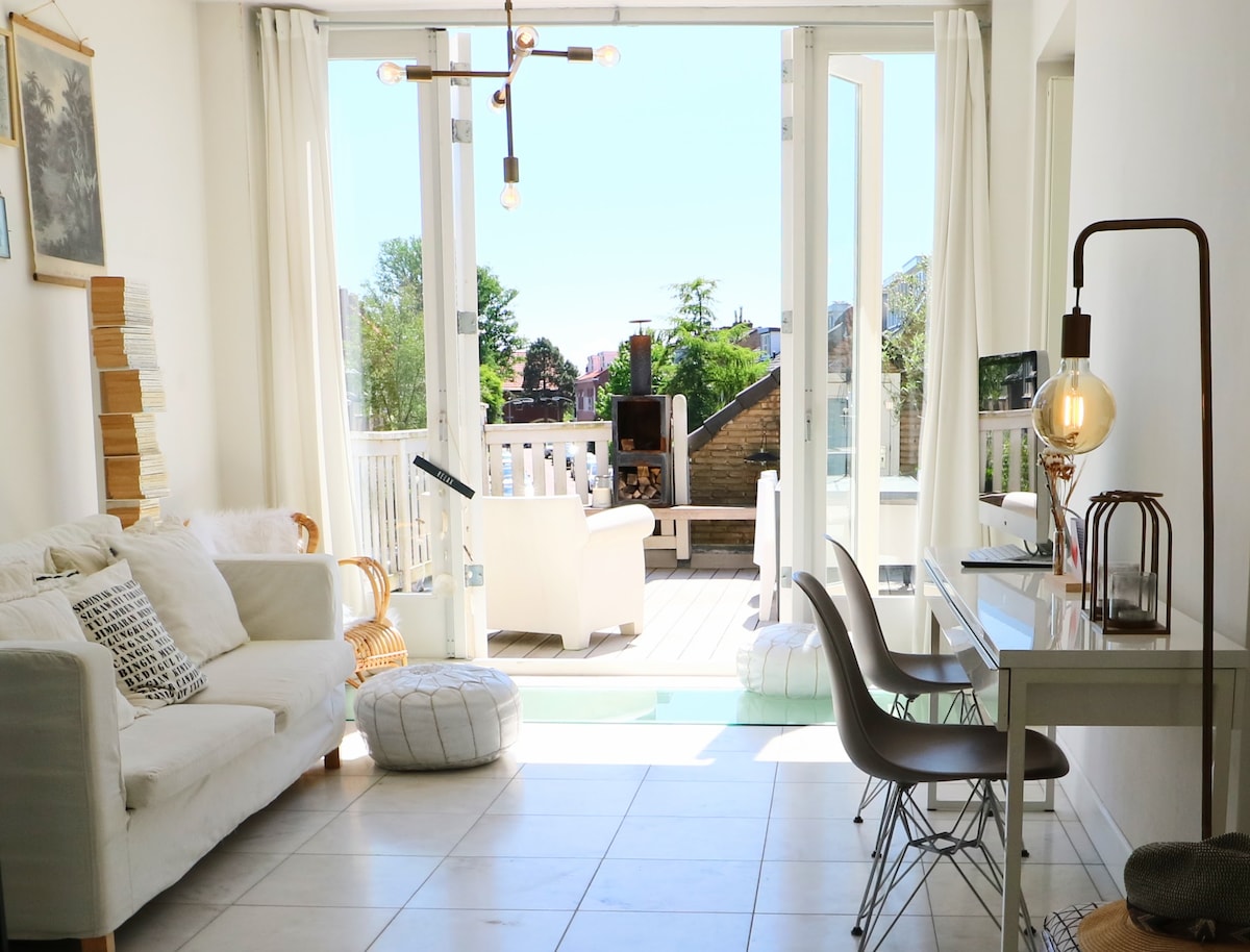 A spacious lounge area features white sofas and a light wooden table, complemented by modern lighting fixtures. Large sliding glass doors open to a bright terrace, enhancing the room's airy ambiance. The design is minimalistic yet inviting, with soft furnishings and natural light.