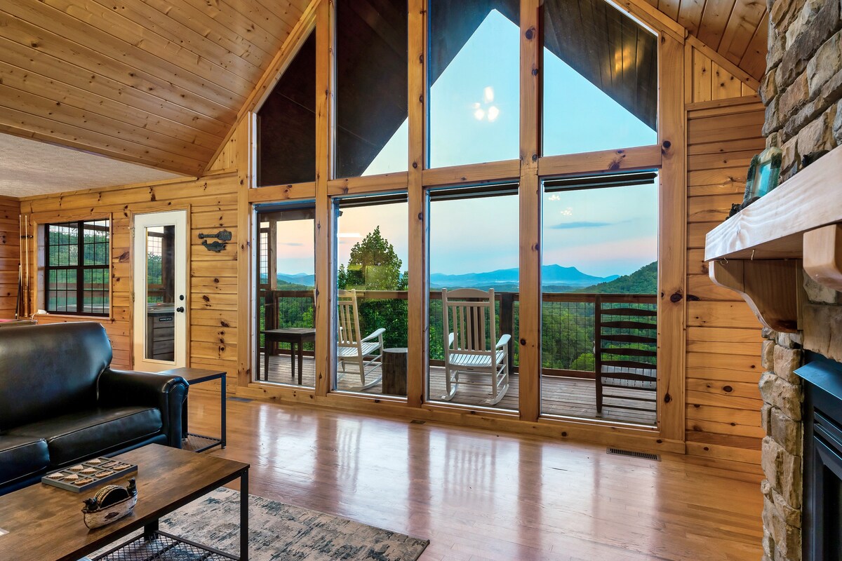 Large windows dominate the living room, offering expansive mountain views. A cozy seating area features a dark sofa and a wooden coffee table. The high ceiling and wooden walls create a warm ambiance, while a door leads to an outside deck with rocking chairs.
