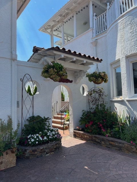 An elegant entrance features a white stucco archway with decorative stonework and lush flower arrangements in hanging planters. Vivid flowers bloom along the path leading to a series of steps, while bright blue skies create a cheerful atmosphere.