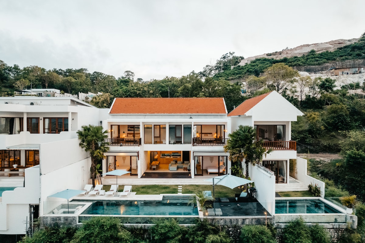 The villa's exterior is showcased, featuring a modern design with multiple balconies. A private infinity pool is visible in the foreground, surrounded by sun loungers and lush greenery. The backdrop includes a gentle hillside, enhancing the serene environment.