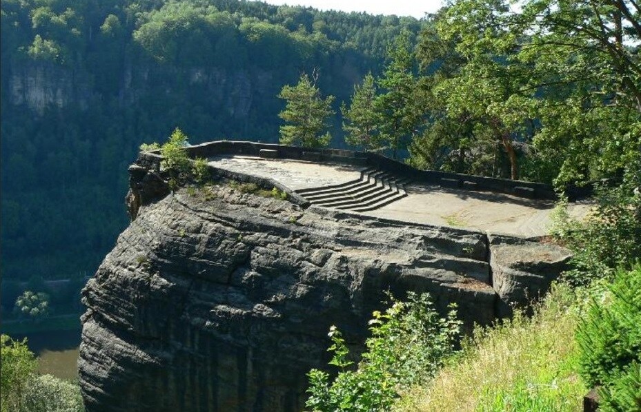 A scenic viewpoint is situated atop a rocky outcrop, with wide stone steps leading down to the edge. Lush greenery surrounds the area, while a river can be seen winding through the valley below, offering a peaceful landscape.