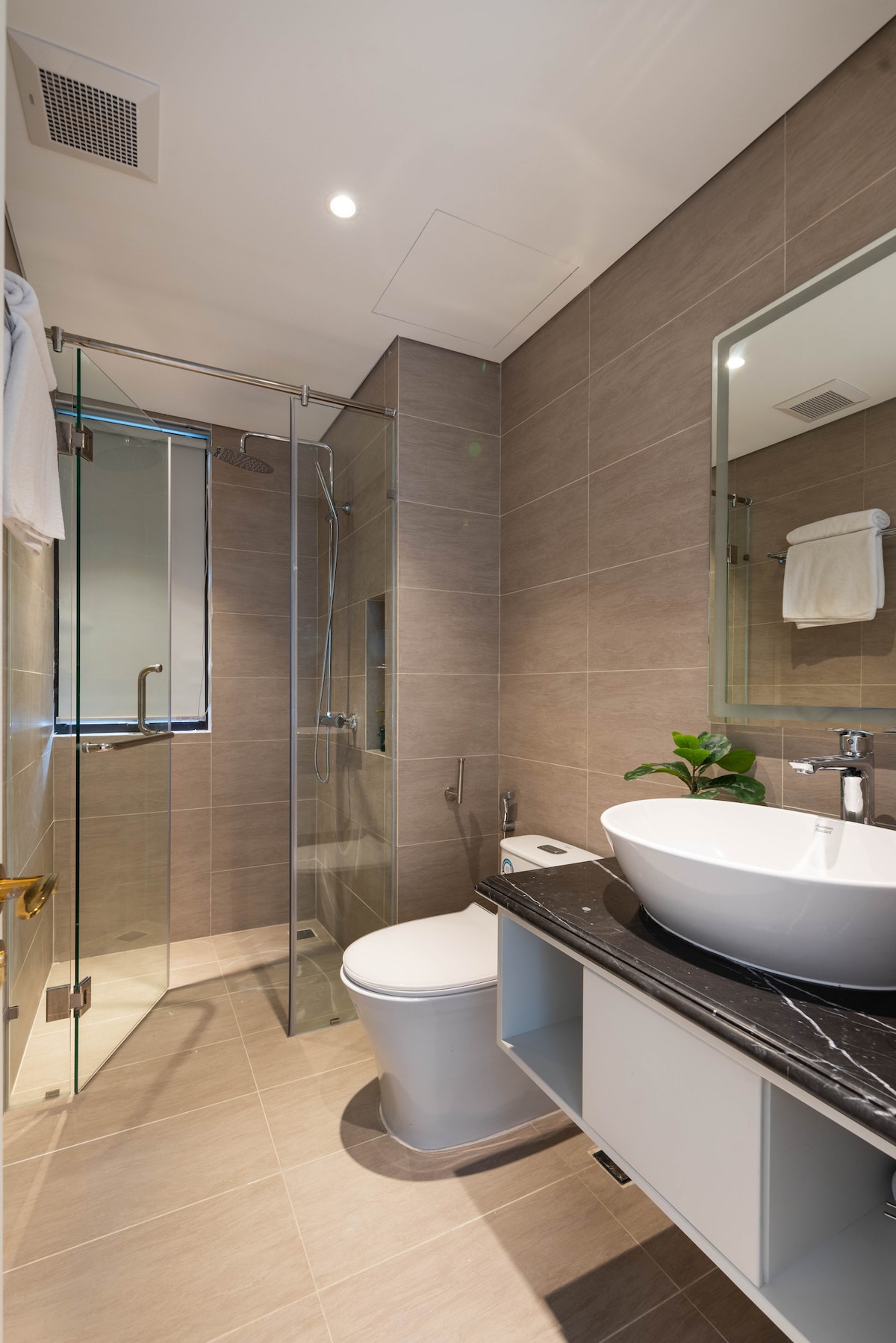 A modern bathroom is showcased, featuring a glass shower enclosure with a rainfall showerhead. A sleek vessel sink rests on a dark marble countertop, accompanied by a wall-mounted mirror. Earthy tiles and gentle lighting create a clean and inviting space.