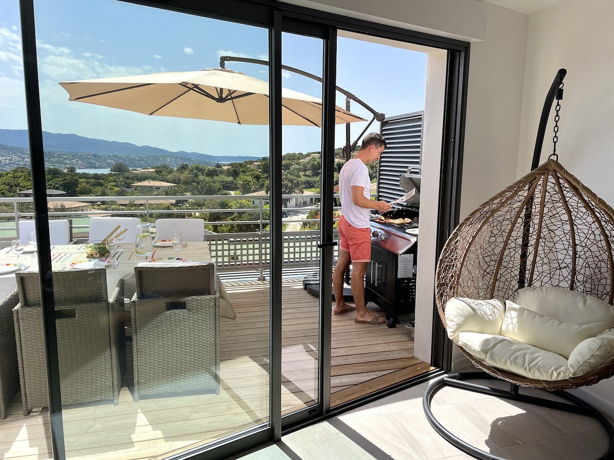 A spacious terrace is featured, showcasing a modern gas barbecue and a stylish hanging chair. The dining area is set with a large table surrounded by several woven chairs. The scene is enhanced by a clear view of the surrounding landscape and a bright blue sky.