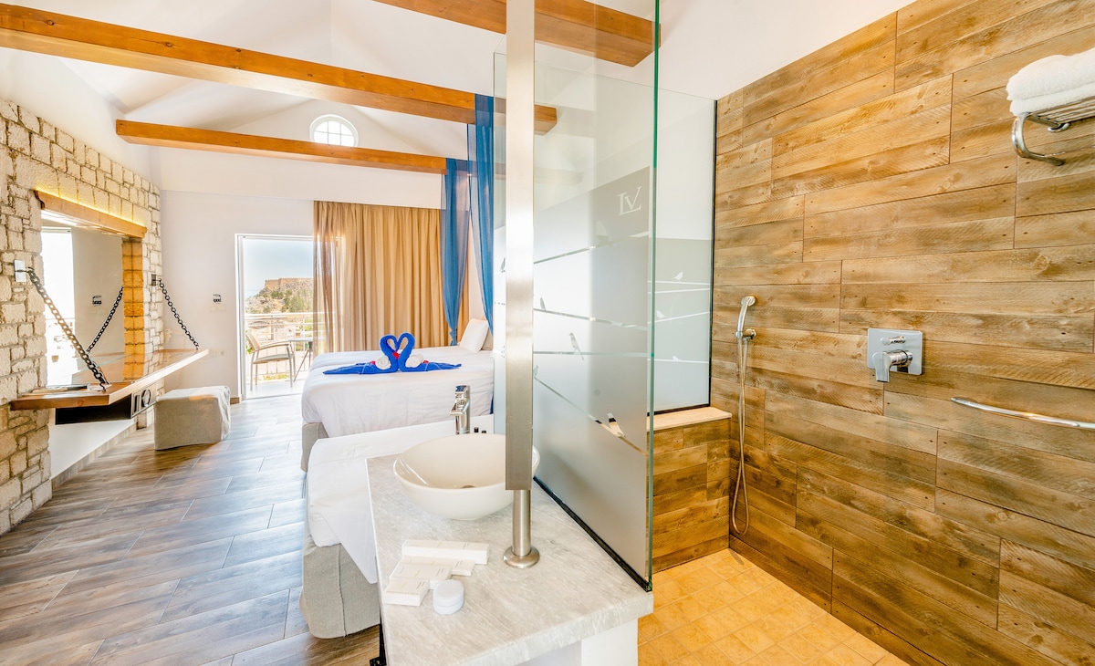 A spacious hotel room features high wooden beams and natural wood accents. A shower area with glass partitions is visible, along with a modern sink on a stone countertop. A balcony opens to beautiful outdoor views, complemented by light curtains and fresh blue decor.