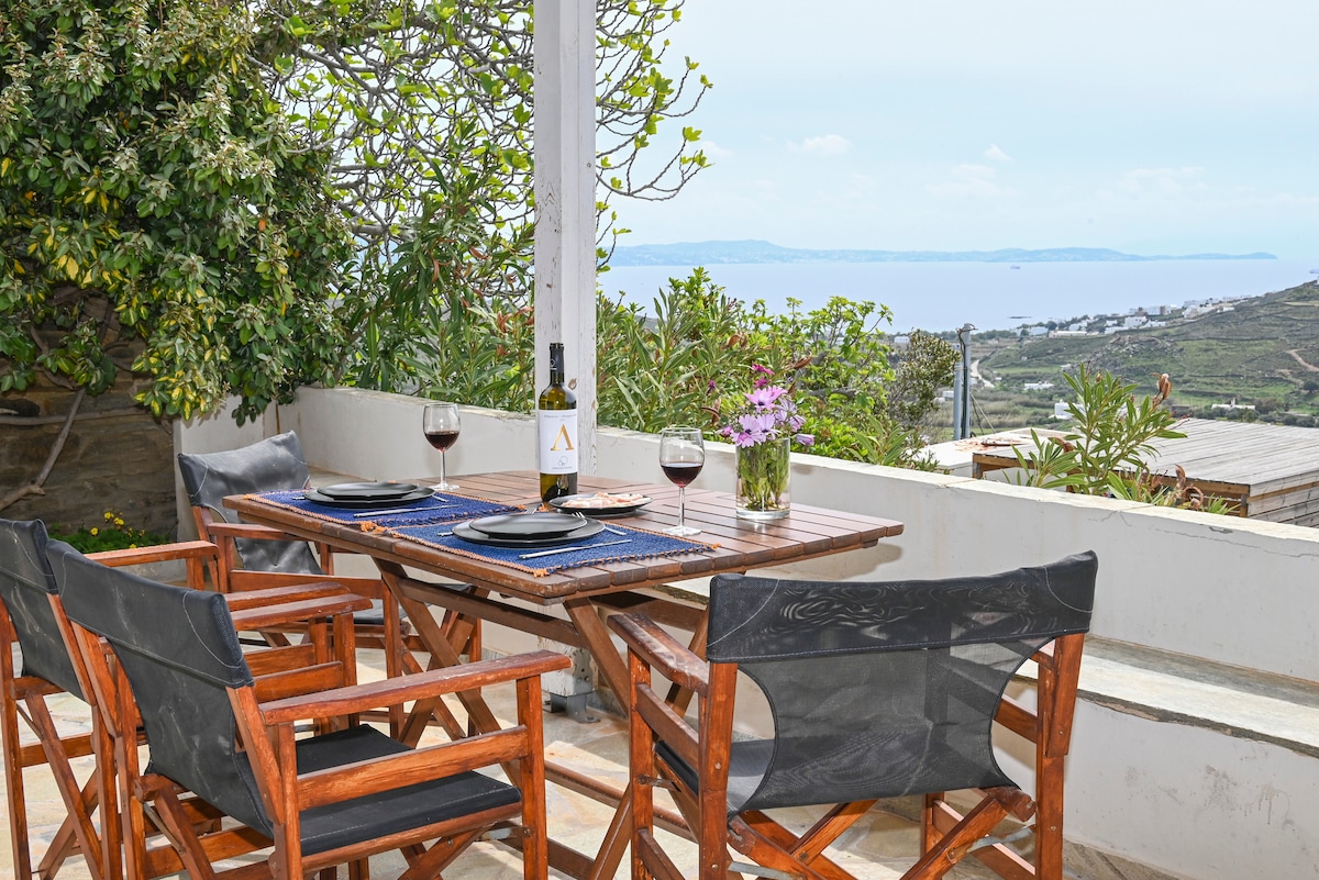 An outdoor dining area is featured, equipped with a wooden table and four folding chairs. A bottle of wine is placed at the center, accompanied by glasses and a vase of flowers. A panoramic view of the sea and surrounding landscape is visible in the background.