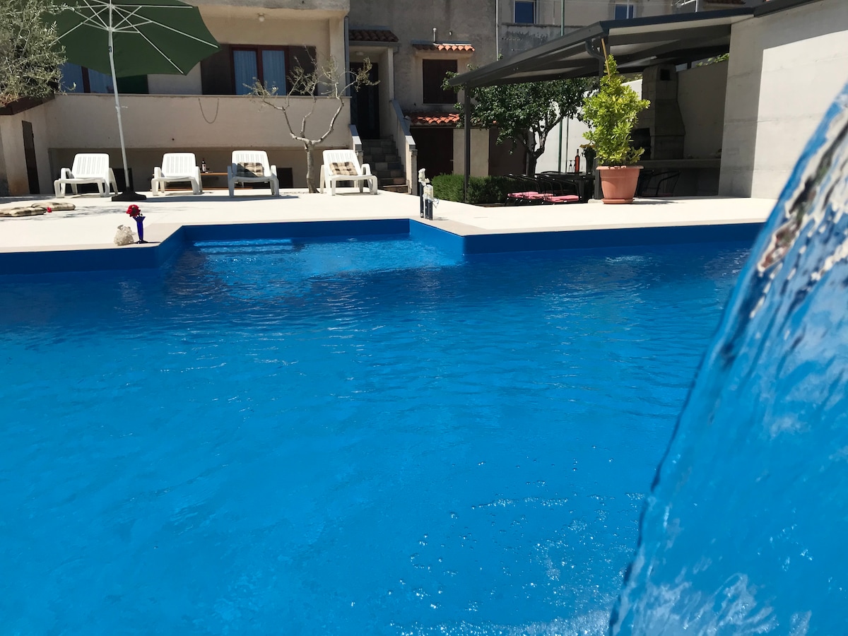 A large swimming pool features a vibrant blue color with a massage waterfall. Sun loungers are arranged around the pool, accompanied by an umbrella. A potted plant is visible near the pool area, providing a touch of greenery.