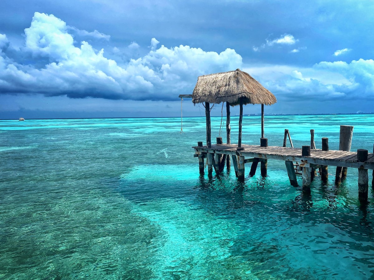 A wooden dock extends over clear turquoise waters, featuring a thatched-roof hut at its end. Dark clouds loom overhead, contrasting with the vibrant blues of the ocean, creating a serene coastal scene.