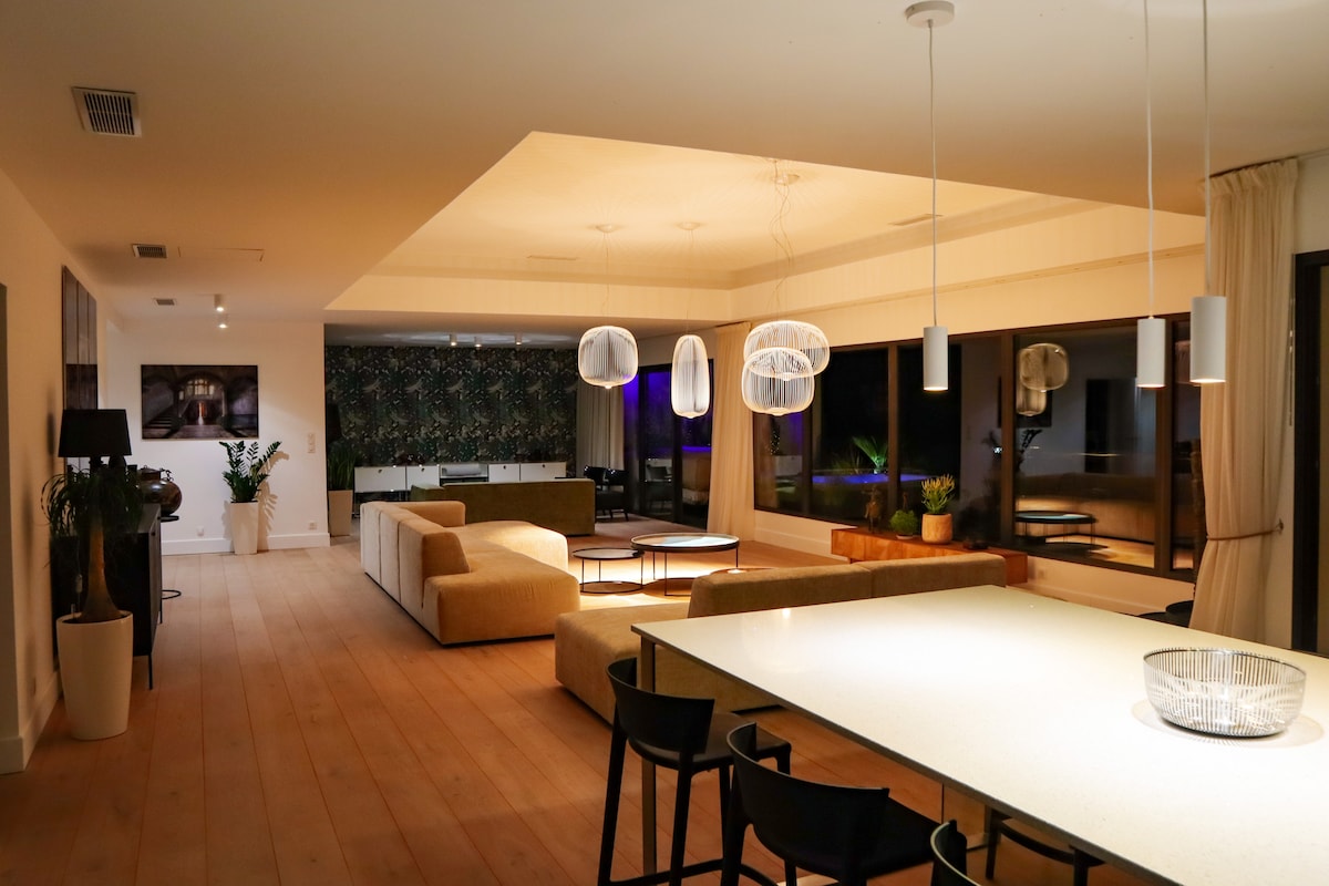 A spacious living area showcases a combination of natural light and stylish decor. Large windows provide views of the outdoor surroundings. The room features modern lighting fixtures, a dining table, and a comfortable seating arrangement with neutral tones, enhancing a contemporary ambiance.