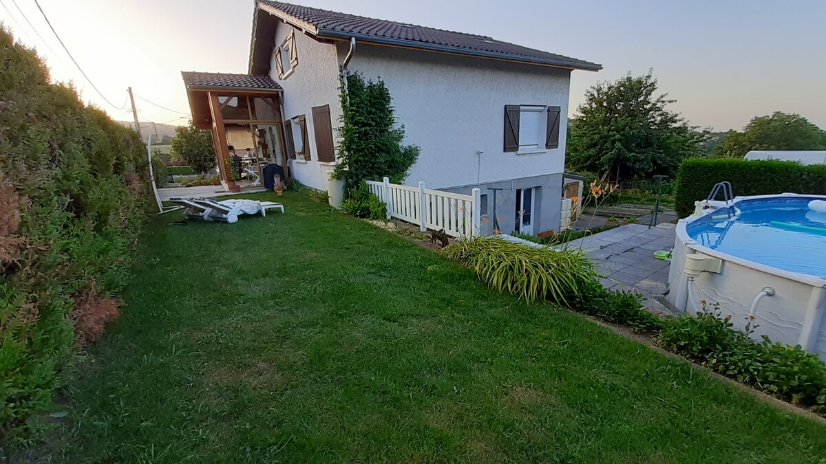 A well-kept garden area features a lush green lawn, bordered by various shrubs. A spacious pool is visible on the right, and a patio area can be seen in front of the house. The warm twilight light enhances the peaceful ambiance of the outdoor space.