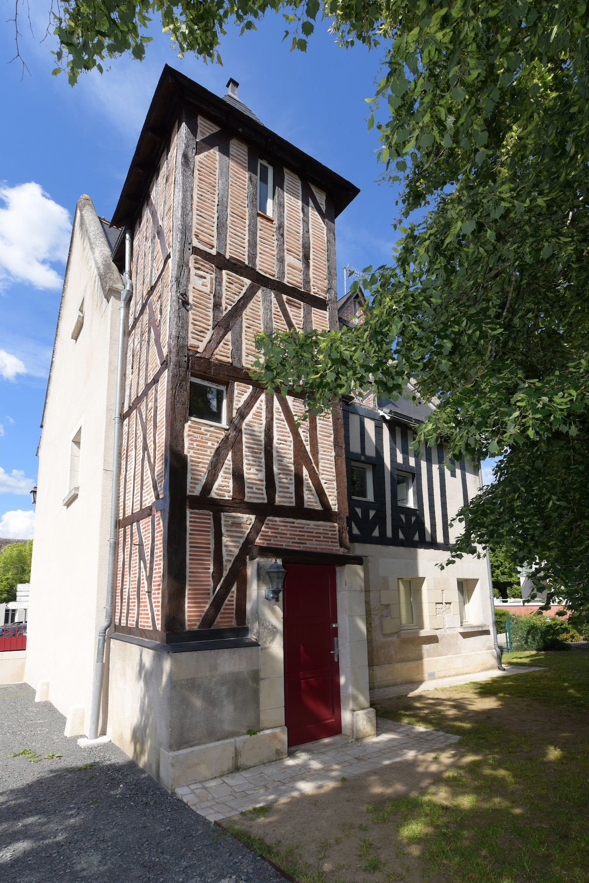 The exterior of a historic building showcases a combination of exposed timber and brickwork, presenting a charming architectural style. A red door serves as the entrance, framed by a stone façade. Lush greenery surrounds the property, adding a natural element to the setting.