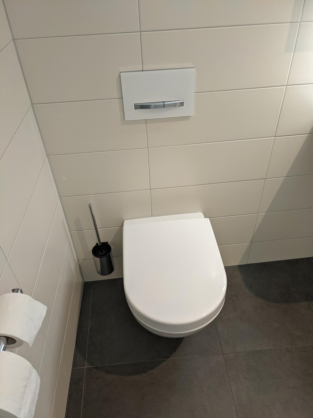A modern bathroom is presented, featuring a wall-mounted toilet with a sleek white finish. The surroundings include neutral-colored tiles with a dark floor. An unobtrusive toilet brush holder is positioned beside the toilet, providing functionality. Two rolls of white toilet paper are neatly arranged on a nearby wall.