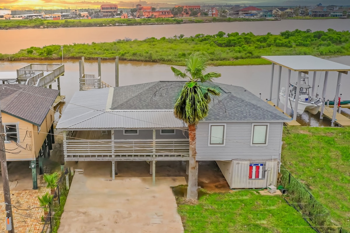 The exterior of a canal front house is shown, featuring a distinct roof and a palm tree in the yard. A spacious driveway allows access, while a fishing dock and boats are visible along the water's edge, with lush greenery in the background.