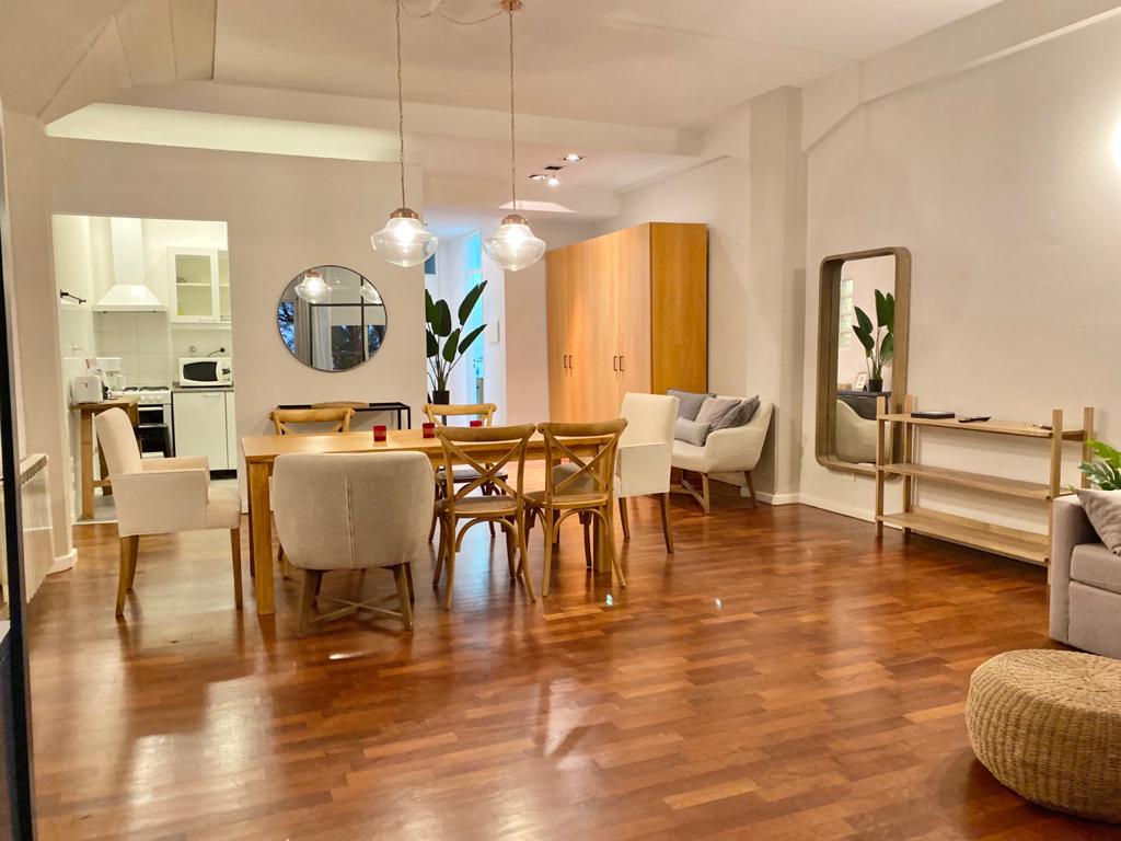 An open living area features polished wooden floors and high ceilings. A large dining table is surrounded by several chairs, while a cozy seating arrangement includes a light-colored sofa and decorative cushions. A round mirror and potted plants add a touch of warmth.