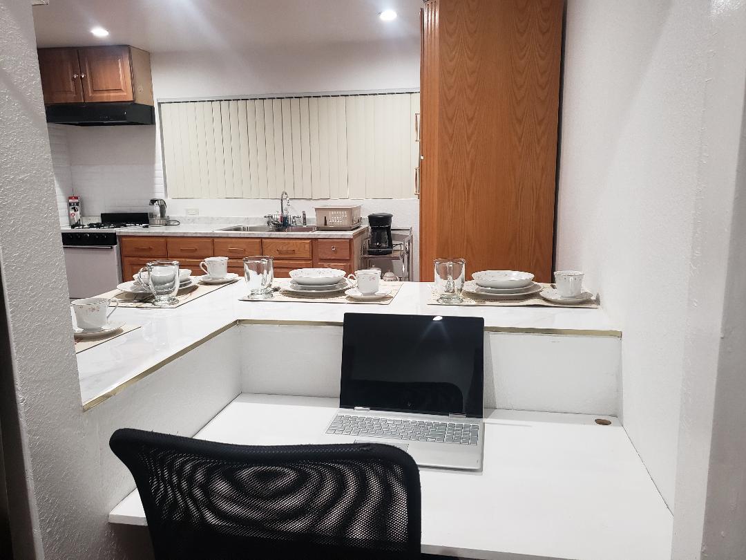 A clean workspace is situated near a kitchenette, featuring a laptop on a simple white desk. The kitchen area includes wooden cabinetry and a well-set dining table with dishes and glassware. Natural light is filtered through horizontal blinds, illuminating the space.