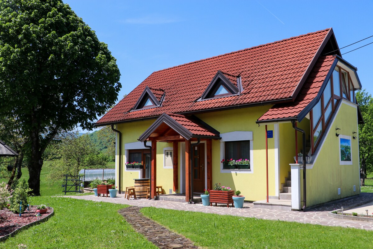 A two-story yellow house features a red roof and decorative gables. Flower boxes adorn the windows, enhancing the home’s charm. A stone pathway leads to the entrance, surrounded by a grassy yard with trees and a view of nature in the background.