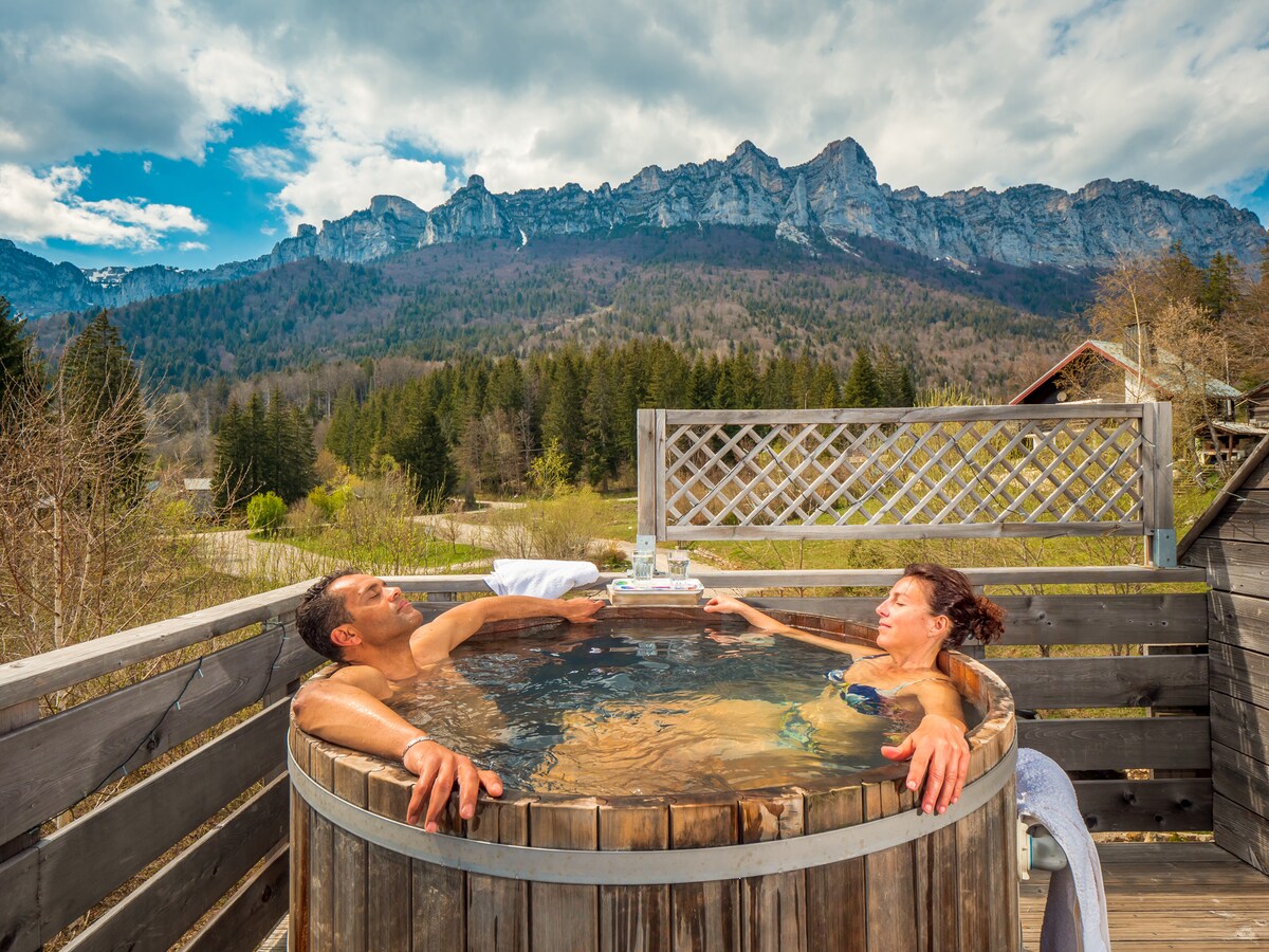 A wooden hot tub is positioned on a terrace, offering panoramic views of distant mountains under a partly cloudy sky. Two individuals are seated inside, enjoying beverages. Surrounding greenery and a wooden lattice provide a natural and serene ambiance.