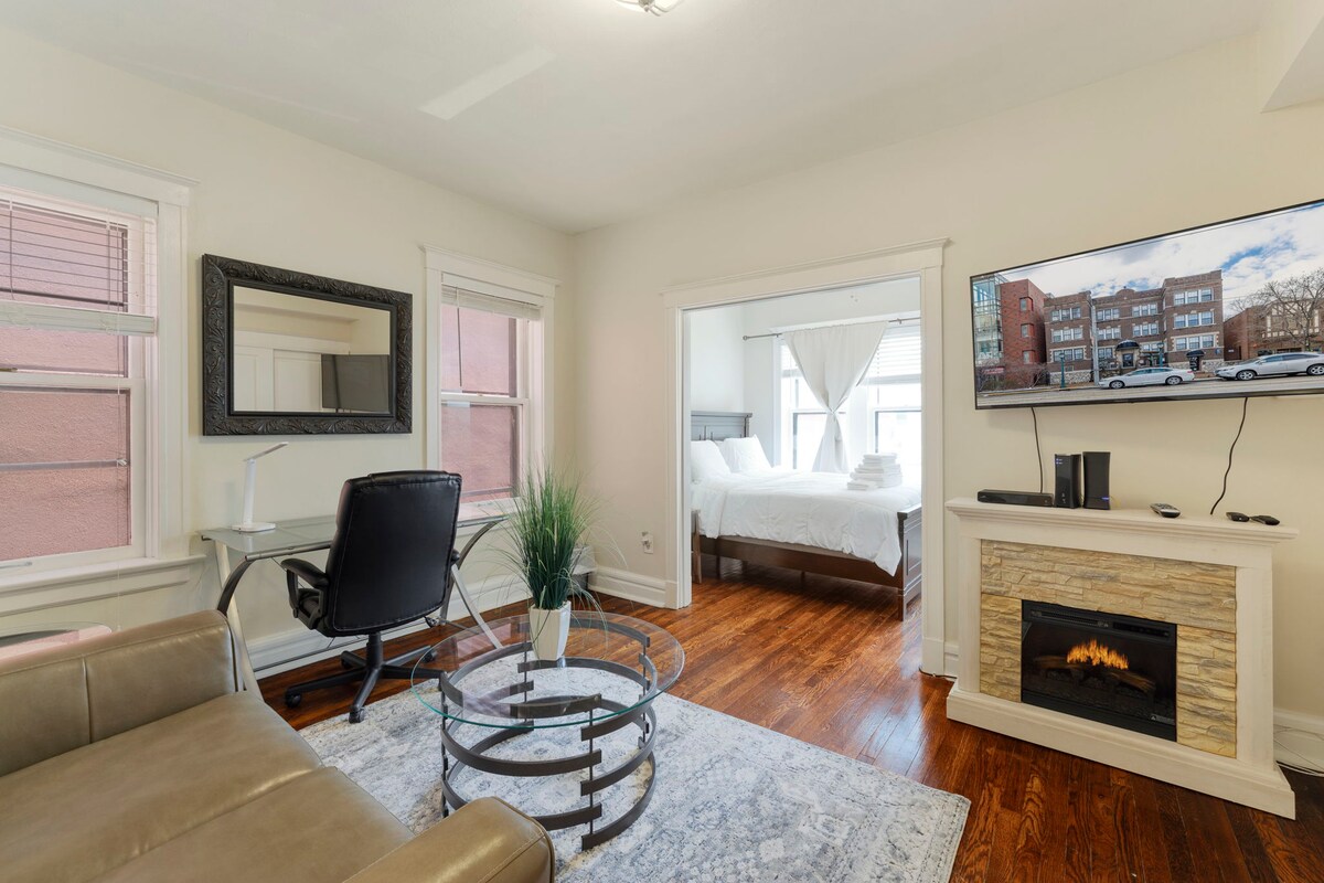 A bright living area features a cozy seating arrangement with a light-colored sofa and a glass coffee table. A workspace is set up with a desk and chair, alongside a flat-screen TV mounted above a stylish electric fireplace. Natural light flows in through large windows.