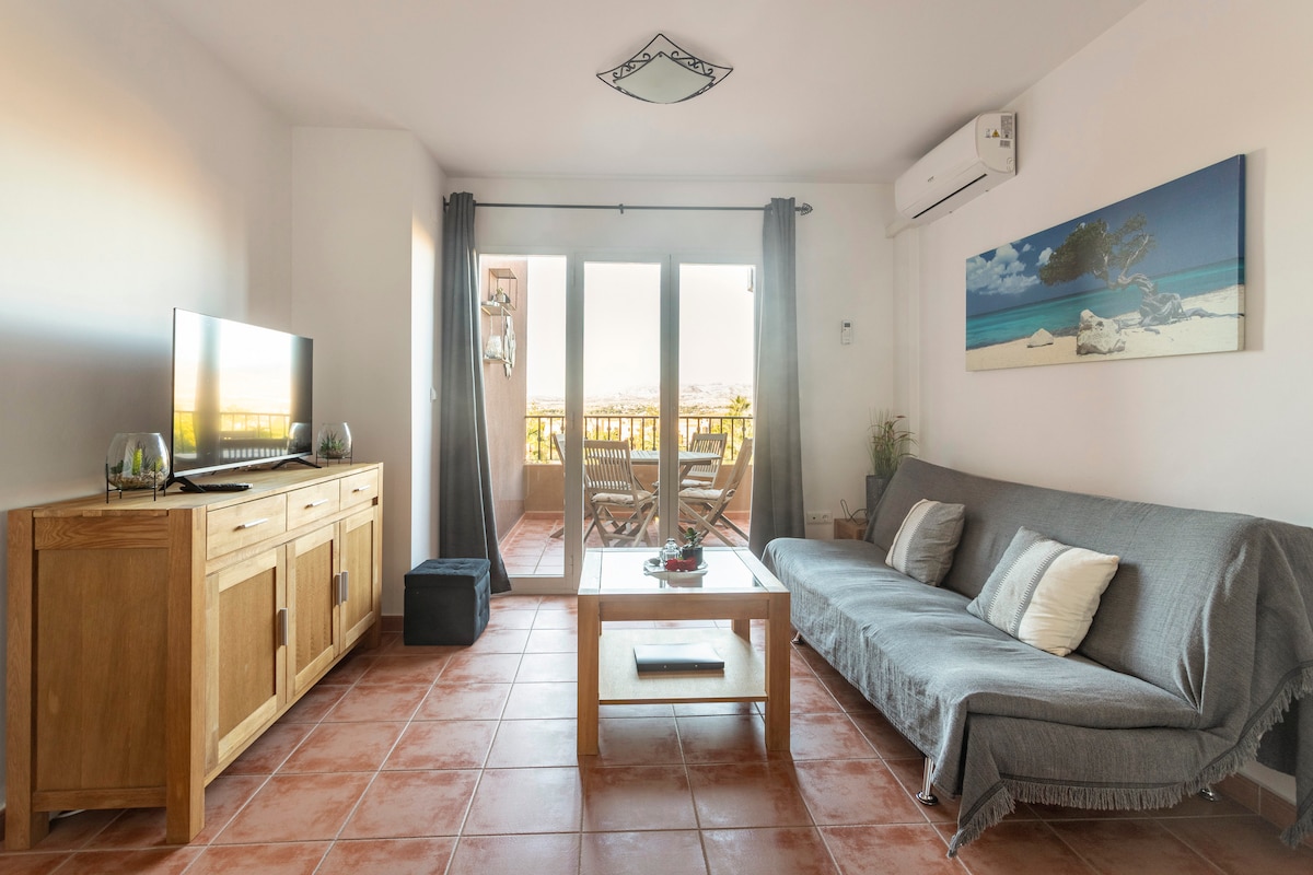 A spacious living area features a comfortable sofa and a wooden coffee table, with a flat-screen TV positioned on a sideboard. Glass doors open to a balcony with an outdoor dining set. Soft natural light fills the room, accentuated by neutral decor.