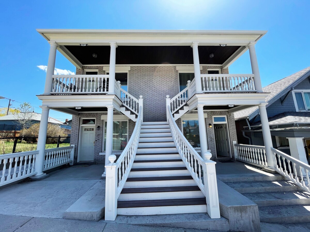 A two-story building features a prominent staircase leading up to a spacious porch with white railings. Pillared balconies adorn both levels, and large windows allow for natural light. The structure's exterior displays a combination of brick and siding, creating a balanced architectural aesthetic.