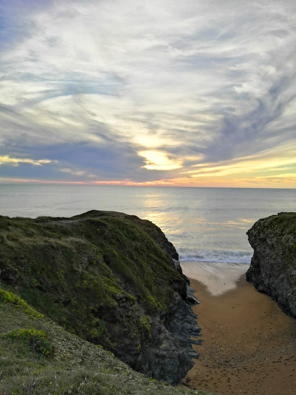 A serene coastal scene is depicted, featuring gentle waves lapping at a sandy beach framed by rocky cliffs. The horizon showcases a sunset with soft hues of orange and purple blending into the sky, providing a natural backdrop of tranquility.