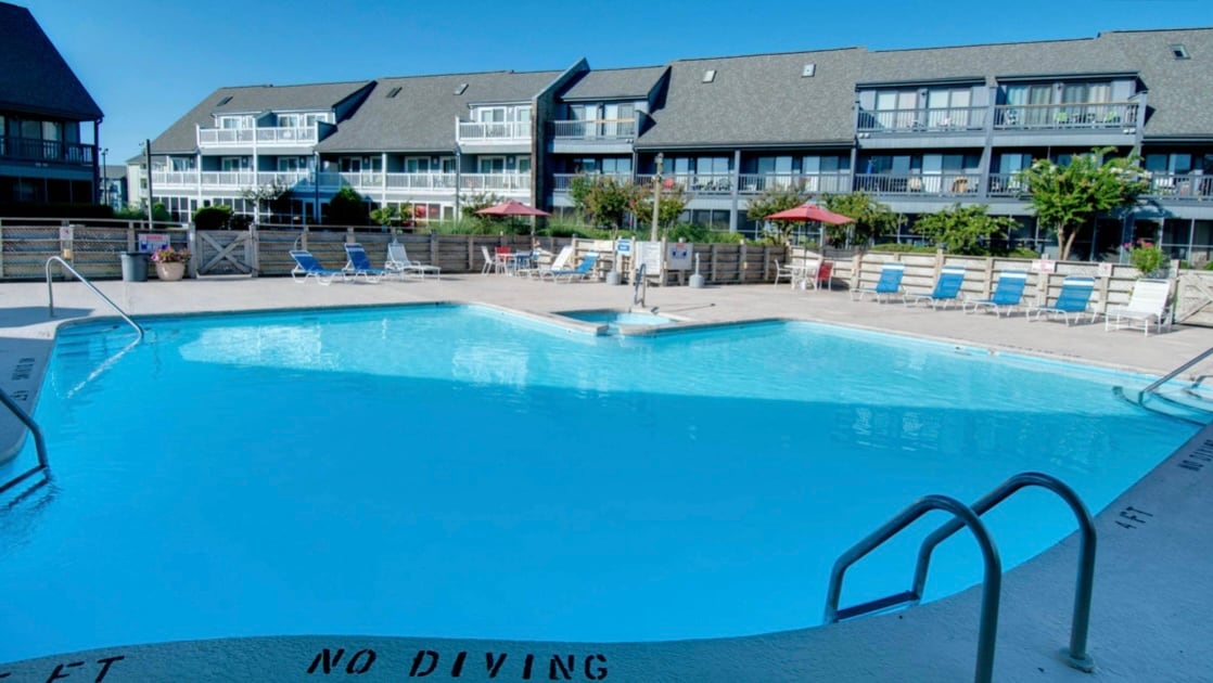 A large outdoor pool is prominently featured in the foreground, surrounded by lounge chairs and umbrellas. Two multi-story buildings are visible in the background, providing shaded balconies overlooking the pool area. Clear blue skies enhance the setting.