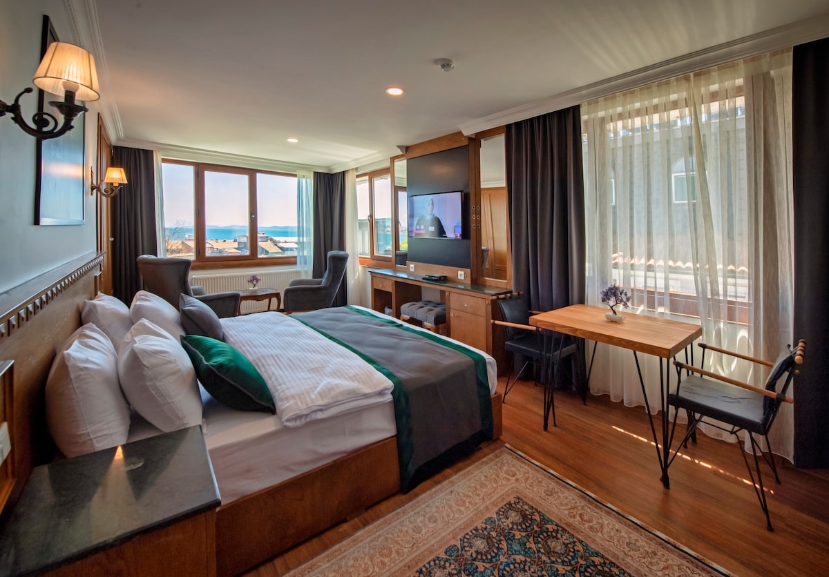 A serene bedroom features a spacious bed with crisp linens, complemented by a wooden accent wall. Two large windows allow natural light to fill the room, revealing a view outside. A small table with chairs and a television on a wooden console enhance the functionality of the space.