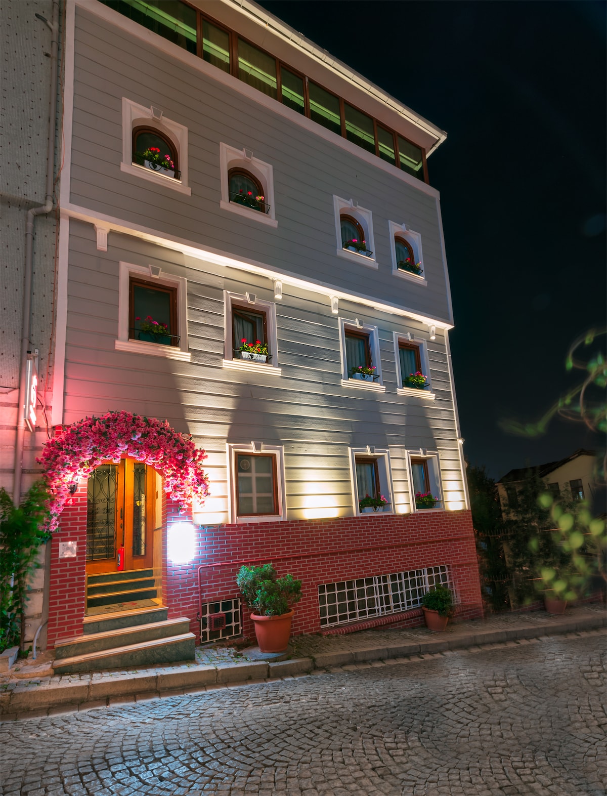 A charming building with a light-colored facade and decorative flower boxes at each window is illuminated at night. The entrance is framed by vibrant pink flowers, with steps leading up to the door. Cobblestone paths create an inviting atmosphere in front of the structure.