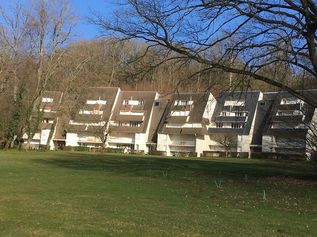 A row of modern residential buildings is set against a backdrop of trees, with gently sloping roofs and large balconies. The vibrant green lawn in front features freshly ground soil, indicating potential landscaping or planting.