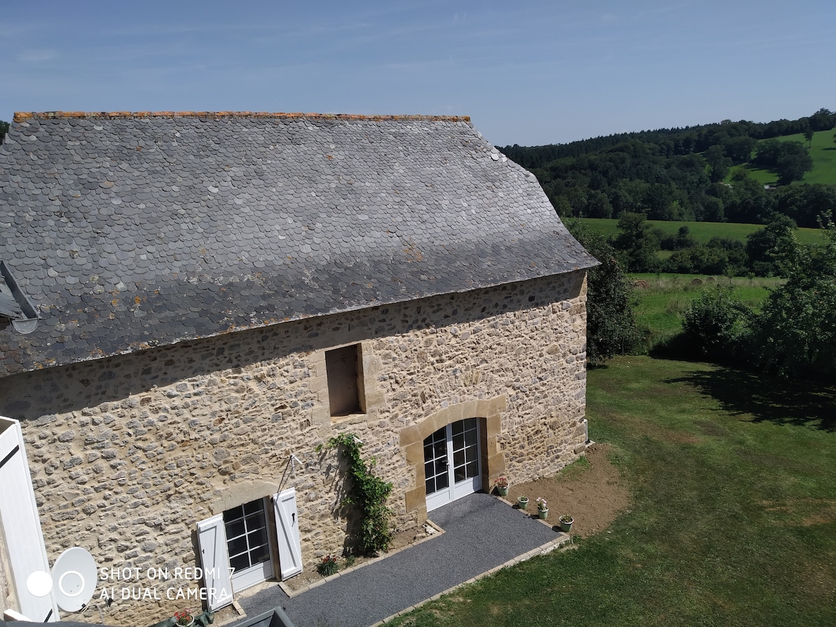 The exterior of a stone farmhouse showcases a slate roof and large windows framed by lush greenery. A gravel path leads to an inviting entryway, with a well-kept lawn extending into the scenic rural landscape beyond.