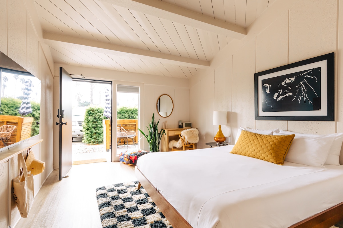 A spacious sleeping area features a large bed with white linens and a mustard yellow decorative pillow. Natural light enters through a door leading to an outdoor space. A round mirror, a stylish lamp, and vibrant artwork complement the minimalist decor.