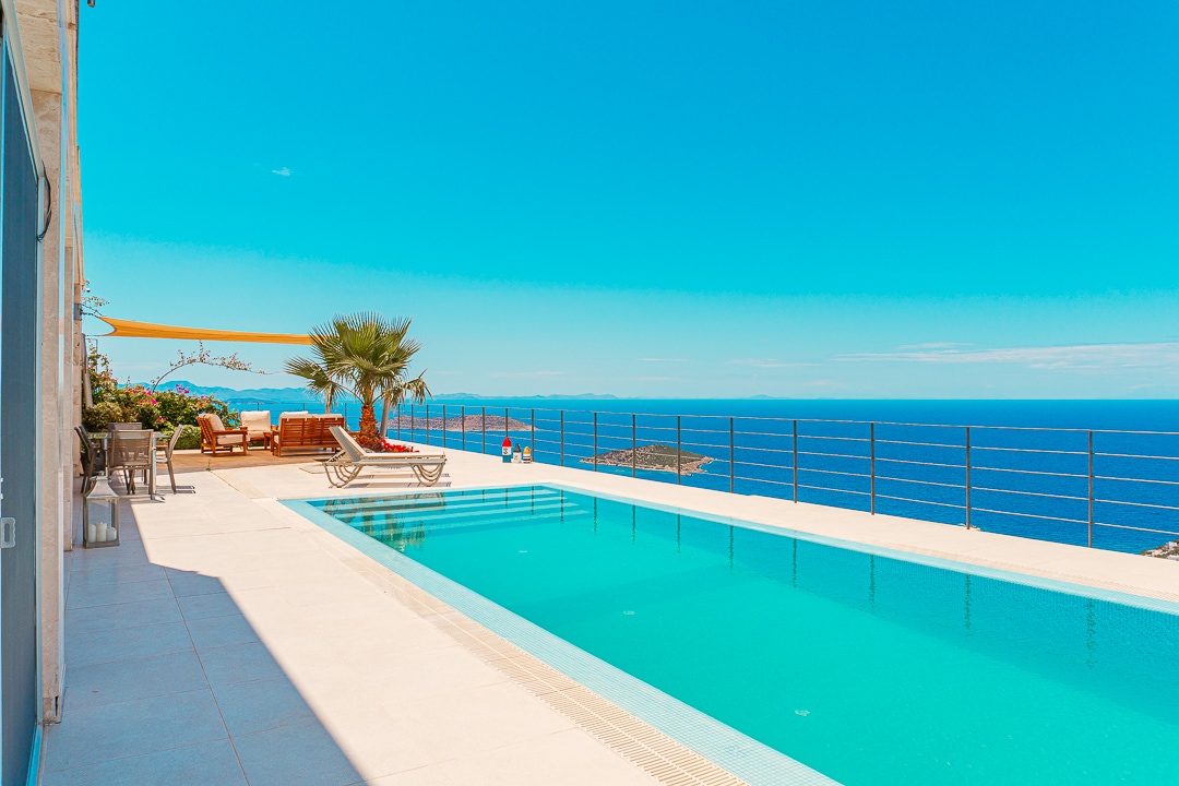 A spacious outdoor area features a sparkling swimming pool with a clear view of the horizon. Sun loungers and a shaded seating area are positioned alongside the pool, complemented by tropical greenery. The scene is framed by bright blue skies and panoramic sea views.