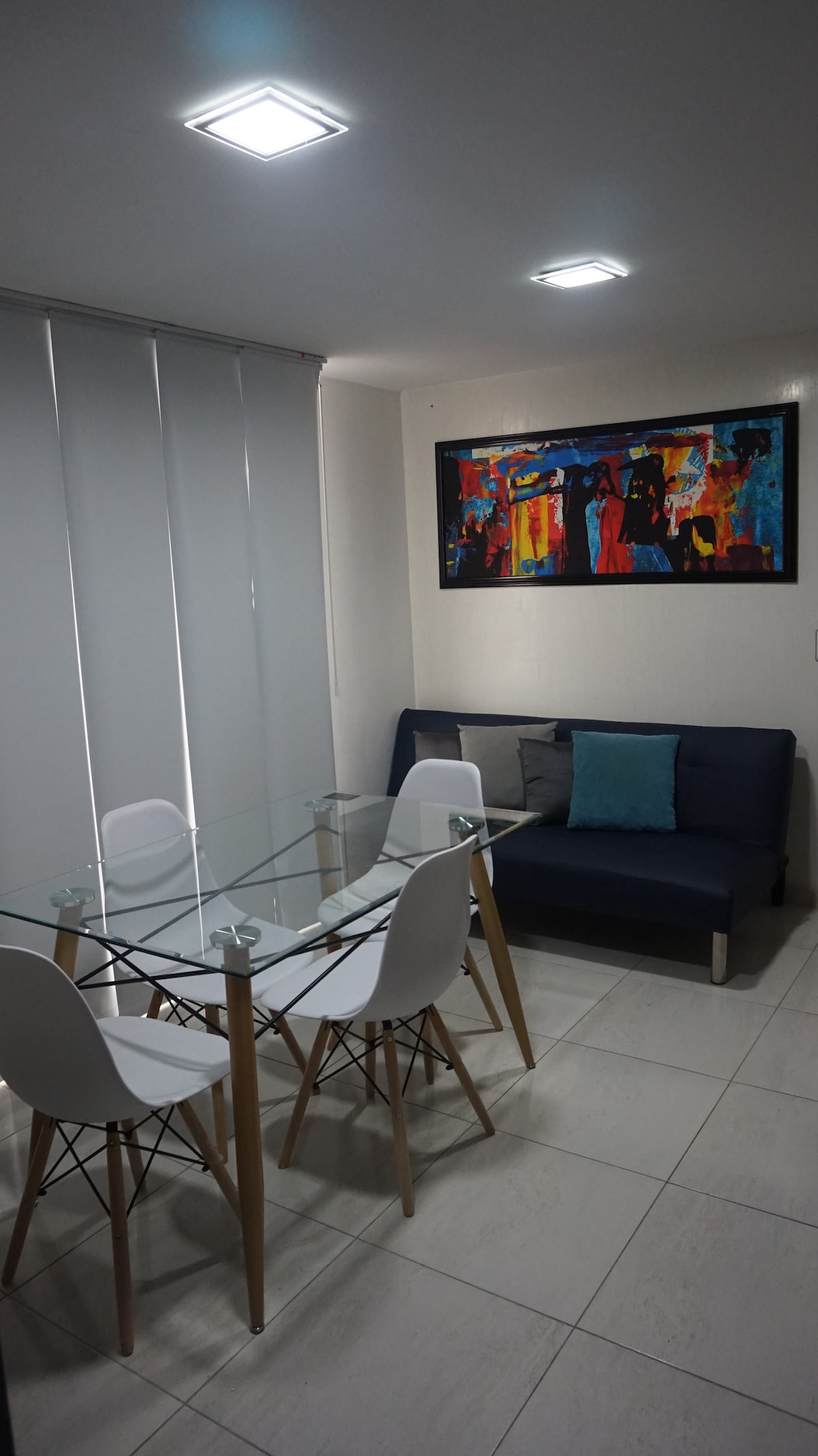 A modern dining area is visible, featuring a glass table surrounded by four white chairs. A navy sofa with a decorative pillow is positioned nearby. A vibrant piece of artwork decorates the wall, with soft lighting from recessed ceiling fixtures enhancing the space.