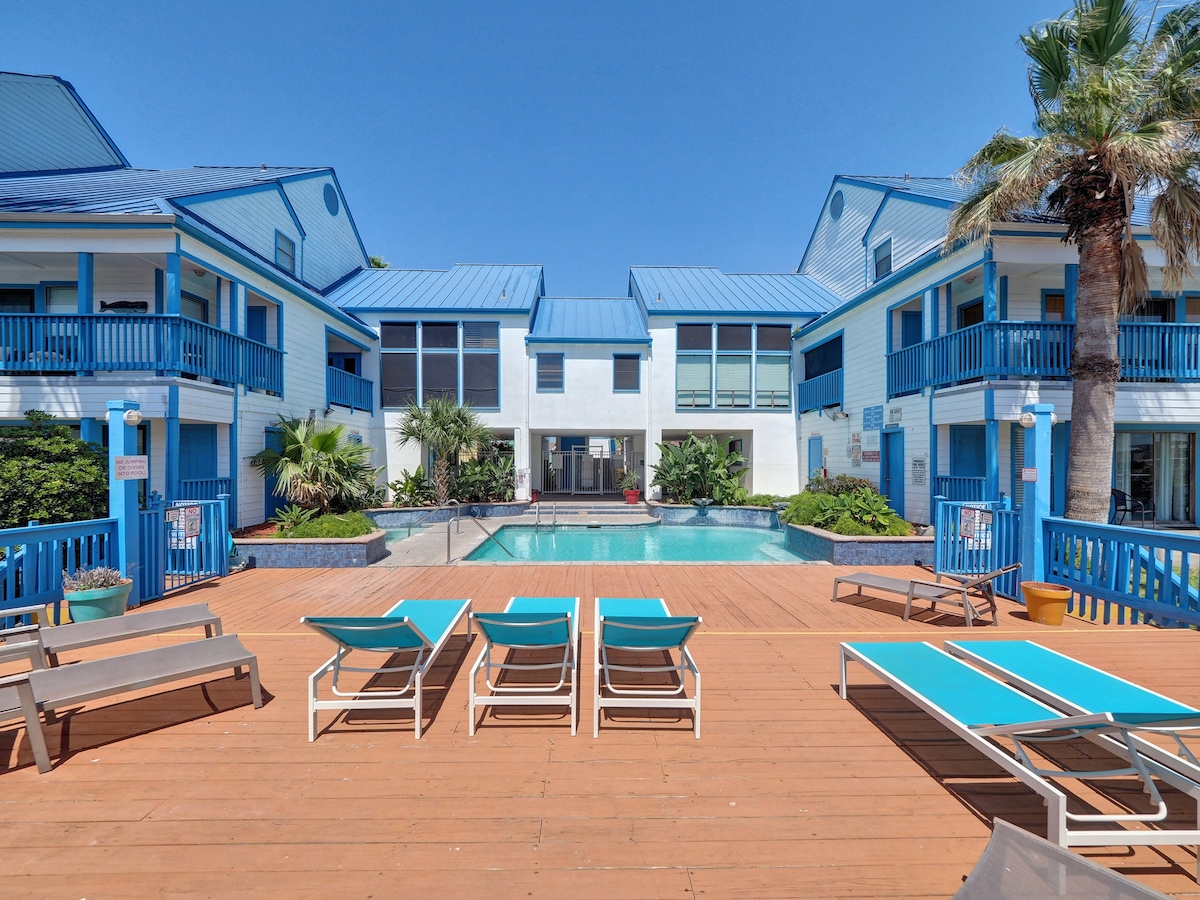 A spacious pool area is framed by two-story buildings painted in light blue, featuring palm trees and lush greenery. Lounge chairs are arranged on a wooden deck, inviting relaxation. Large windows provide a view into the adjacent interior spaces.