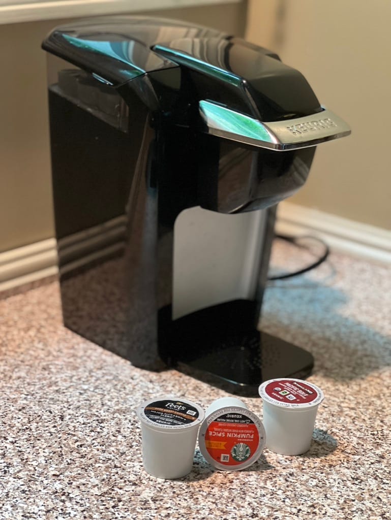 A coffee maker is positioned on a textured countertop, beside three coffee pods. The sleek, black appliance features a modern design, suitable for single-serve brewing. Light is reflected off its surface, highlighting its polished finish.