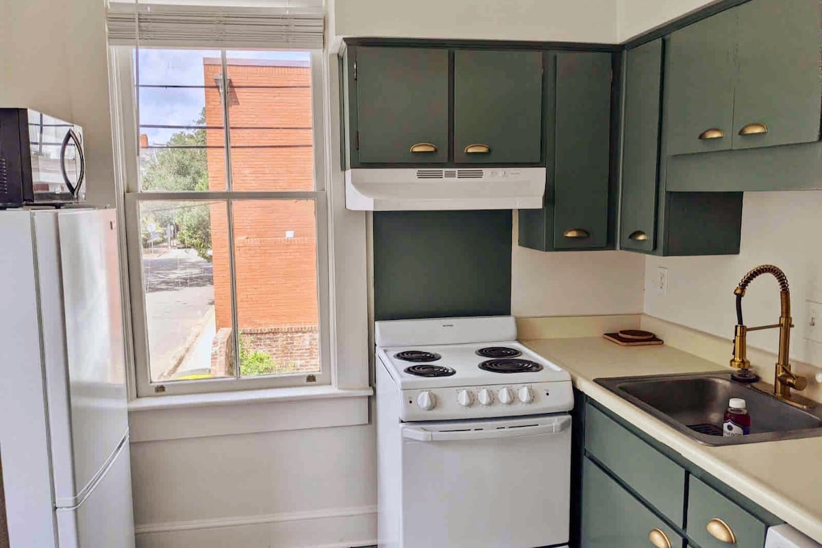 The kitchen features a combination of green cabinetry and a light countertop. An electric stove and oven are present, alongside a sink with a gold-colored faucet. Natural light fills the space through a nearby window, providing a view of the outside street.