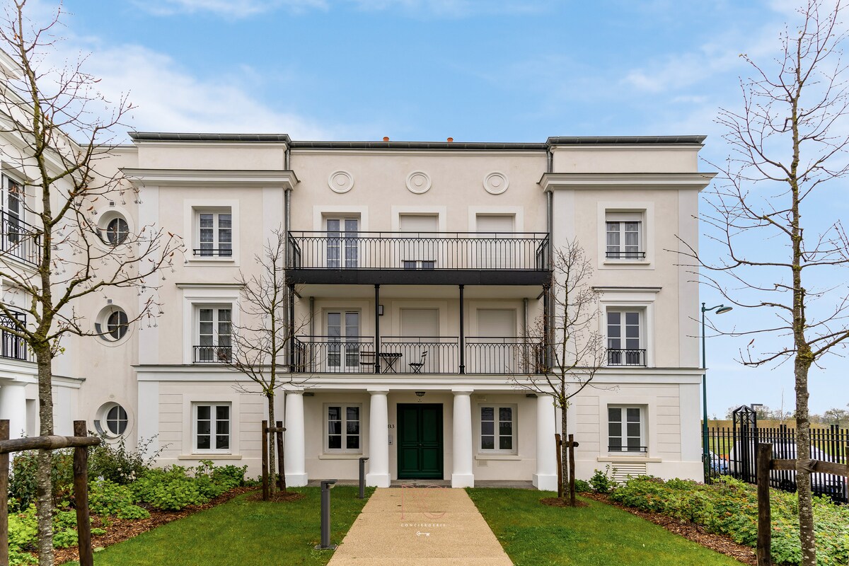 The modern building features a clean, white exterior with multiple windows providing natural light. A welcoming entrance is highlighted by a green door, complemented by a balcony on the first floor. Surrounding the property, neatly trimmed trees and greenery enhance the overall setting.