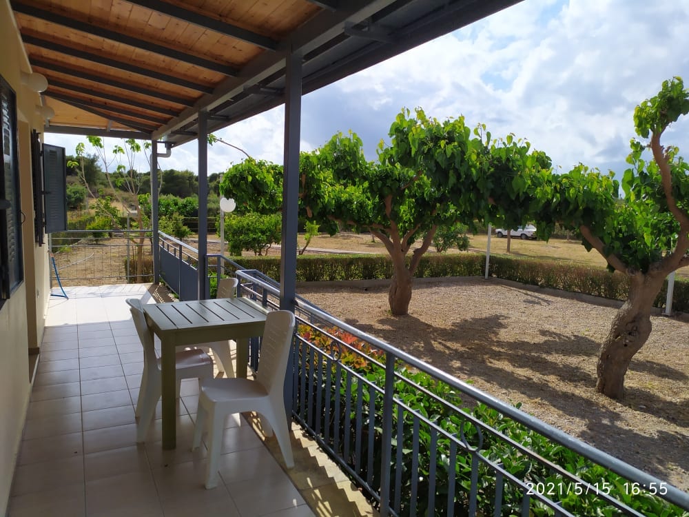 A shaded patio area is featured with a simple outdoor table and a chair. A view of well-maintained greenery and trees is visible in the background, providing a serene outdoor space for relaxation.