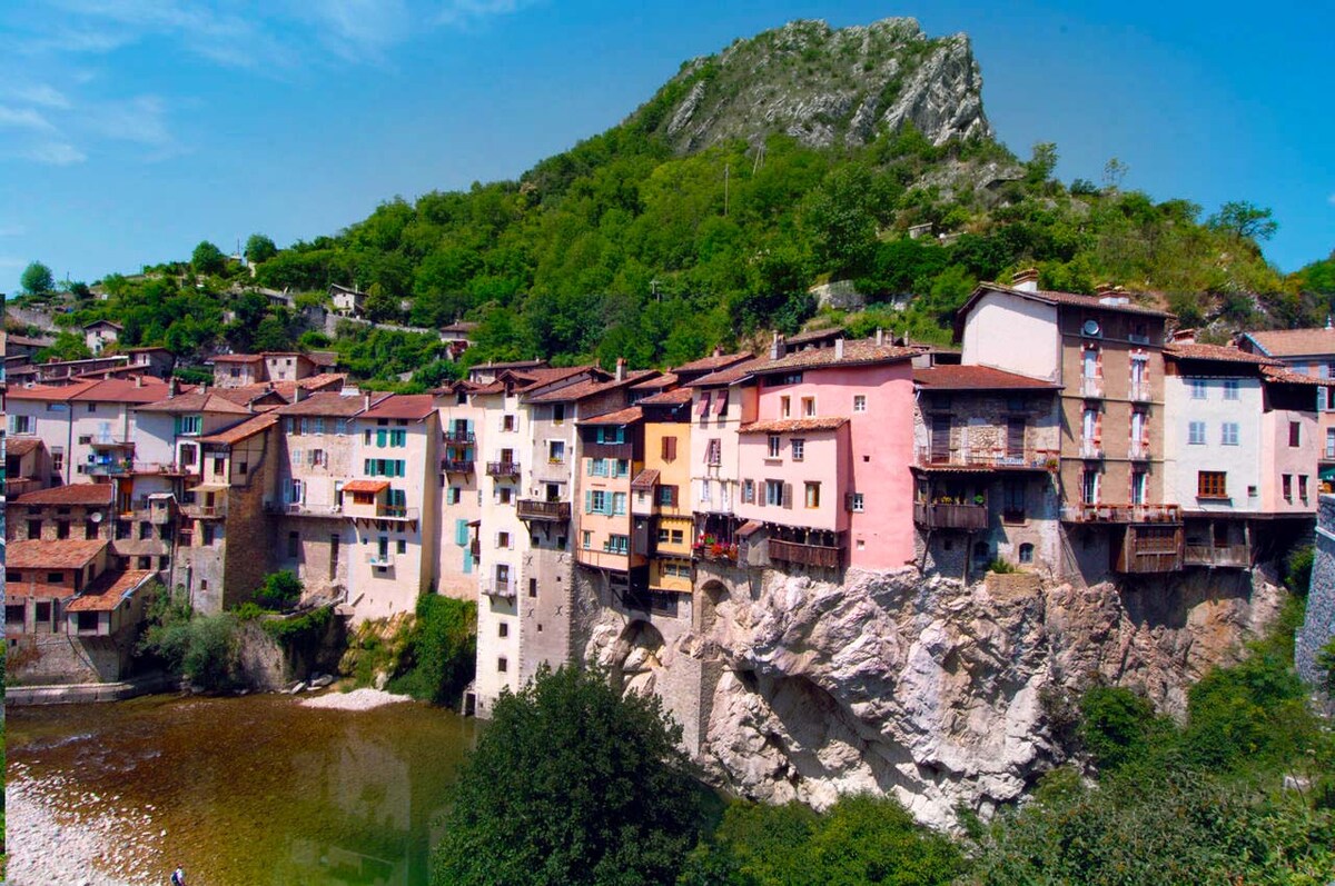 A charming village is nestled against a rocky hillside, showcasing a mix of pastel-colored buildings that cling to the slopes. The scene is framed by lush greenery, with a calm river flowing in front, reflecting the natural beauty of the surroundings.