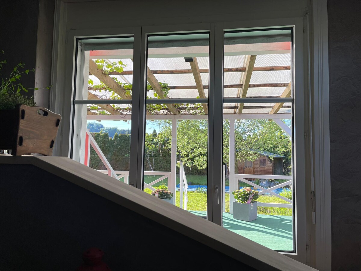 A view through large glass doors reveals a covered terrace with wooden beams, surrounded by greenery. Potted flowers are visible on the terrace floor, and an expansive garden stretches out beyond, enhancing the serene outdoor environment.