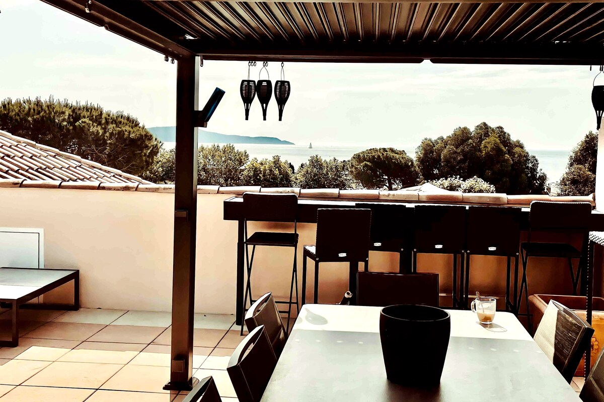 An outdoor rooftop terrace is showcased, featuring a table surrounded by chairs and a pergola overhead. Coastal views are seen in the background, highlighting greenery and the distant sea. Light colors dominate the decor, enhancing the open and airy atmosphere.