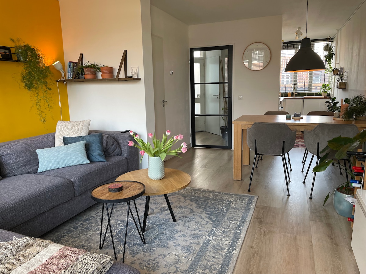 A modern living space is presented, featuring a gray sofa adorned with throw pillows. Two round wooden tables sit atop a patterned rug, while a bouquet of tulips adds a touch of color. The dining area includes a wooden table and chairs, with natural light filtering through a nearby window.