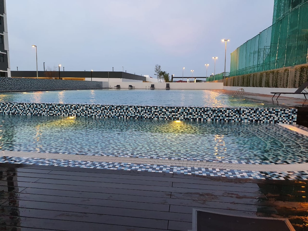 The outdoor swimming pool is bordered by sleek wooden decking and features a mosaic tile pattern. Soft lighting illuminates the water, creating a calming atmosphere. Lounge chairs are positioned around the pool, and a section for swimming is visible in the background.