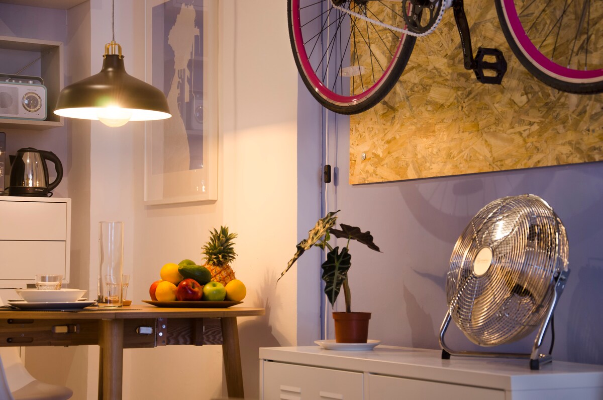 A cozy dining area is highlighted by a round wooden table set with dinnerware and a pitcher of water. A stylish ceiling light illuminates the space, while a colorful bicycle hangs on the wall beside a fan and a small potted plant among fresh fruits.