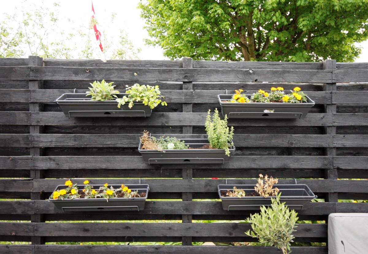 A wooden fence in a dark finish displays several vertical planters. The planters contain a variety of greenery and vibrant yellow flowers, adding a touch of natural beauty to the outdoor space. Lush foliage can be seen in the background, enhancing the serene environment.