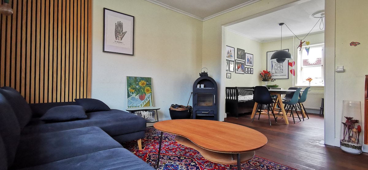 A cozy lounge area features a dark sofa accompanied by a circular wooden coffee table resting on a vibrant area rug. Natural light fills the room, highlighting framed artwork on the walls and a view of the dining area through an open doorway.
