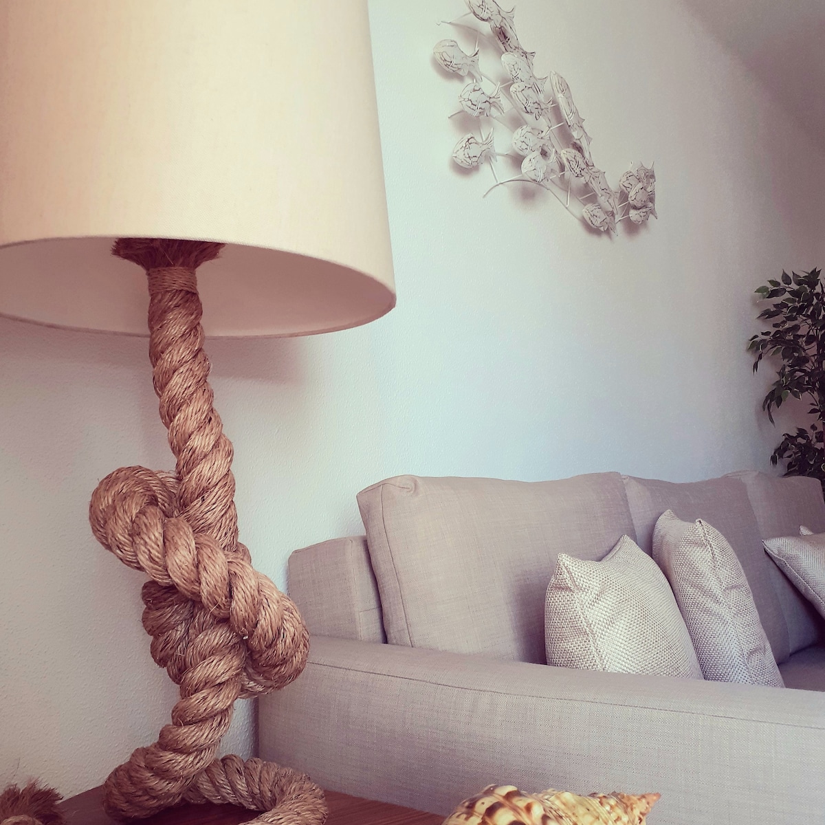 A lamp with a rope-inspired base is positioned beside a light-colored sofa adorned with decorative cushions. A subtle wall decoration featuring floral motifs is displayed in the background. Natural elements, such as seashells, are present on a wooden table.