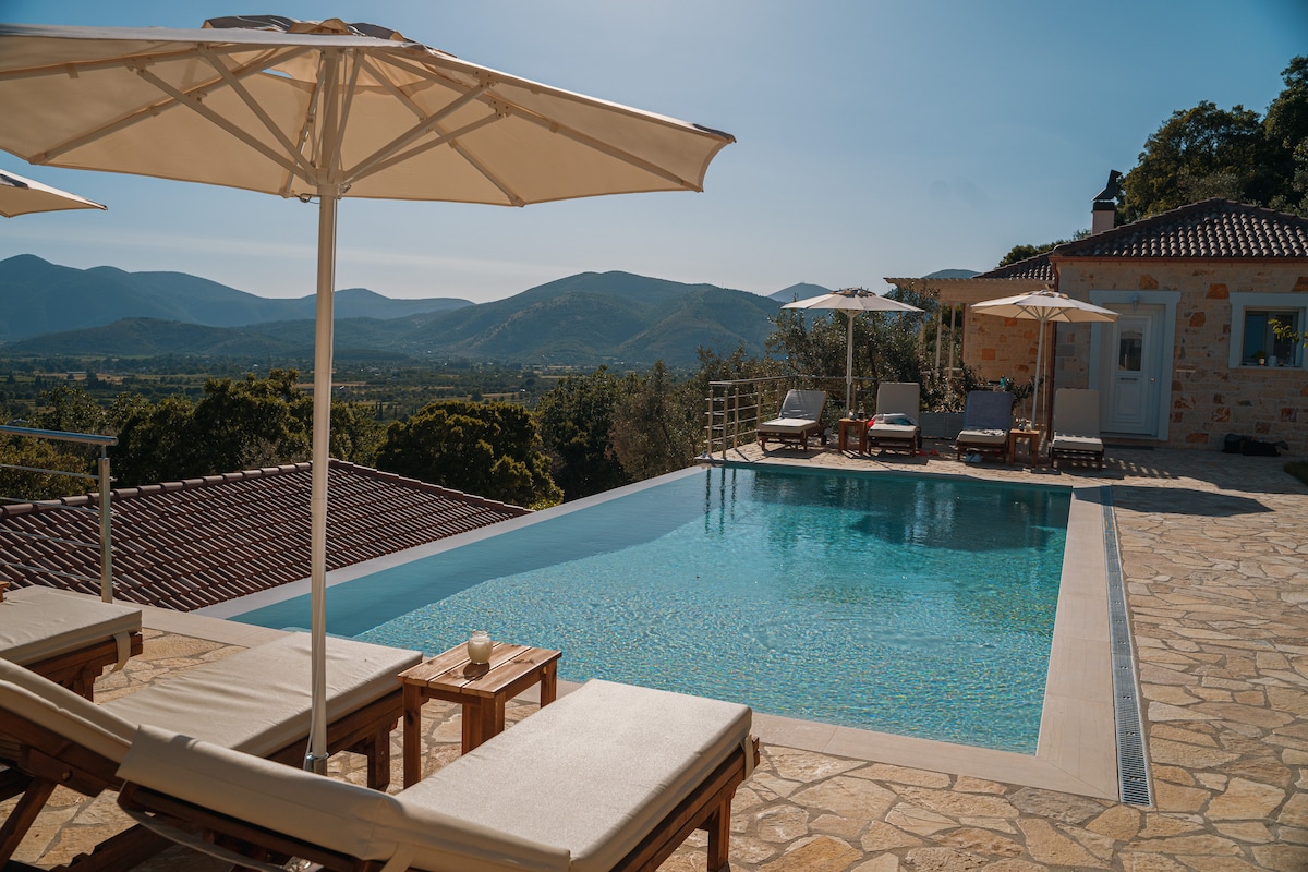 An infinity pool with clear blue water is bordered by padded sunbeds and sunshade umbrellas. The view extends to rolling hills and mountains in the background, under a bright blue sky. The pool area is framed by natural stone flooring, offering a relaxing outdoor space.